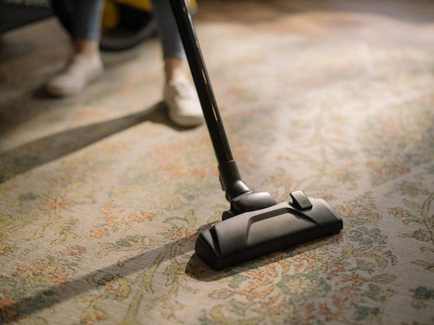 A person in casual clothing, kneeling on a patterned area rug, engaged in surface cleaning with a bright yellow vacuum cleaner equipped with a flexible hose. The individual is using a cloth to wipe th