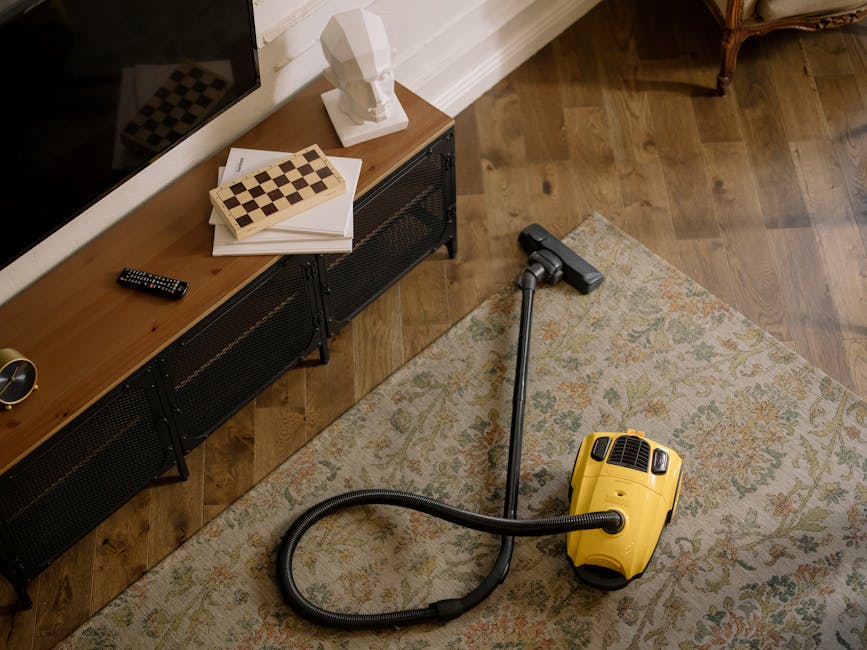 A person kneeling on a patterned carpet in a room with wooden furniture, using a yellow portable vacuum cleaner with a black hose to perform surface cleaning. The individual is adjusting the vacuum's 